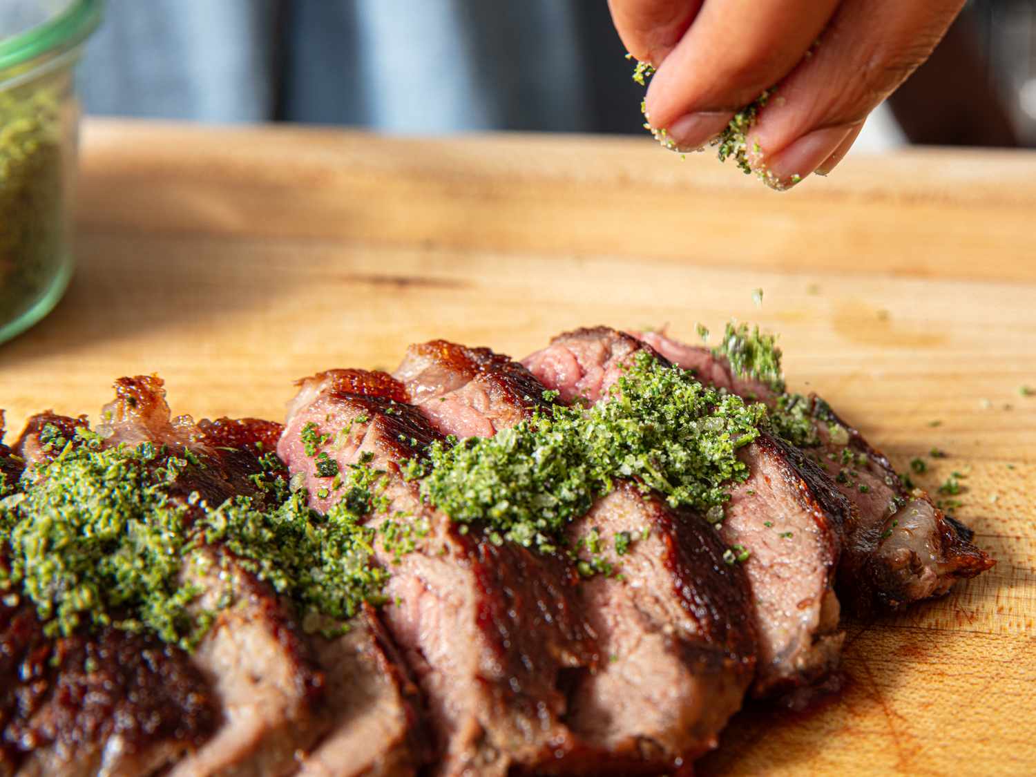 A closeup of sliced steak on a wooden surface being sprinkled with herb seasoning by a persons hand