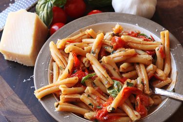Finished dish of pasta with blistered cherry tomato sauce, next to a block of cheese, basil sprig, garlic, and tomatoes