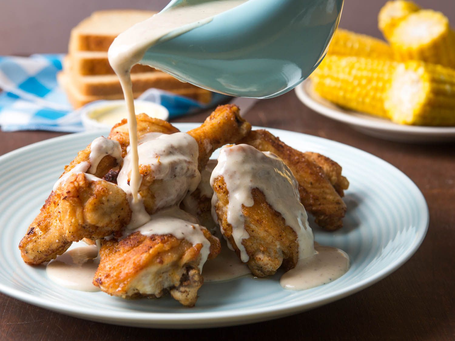 Pouring white gravy over a plate of Maryland fried chicken with corn and bread in the background.