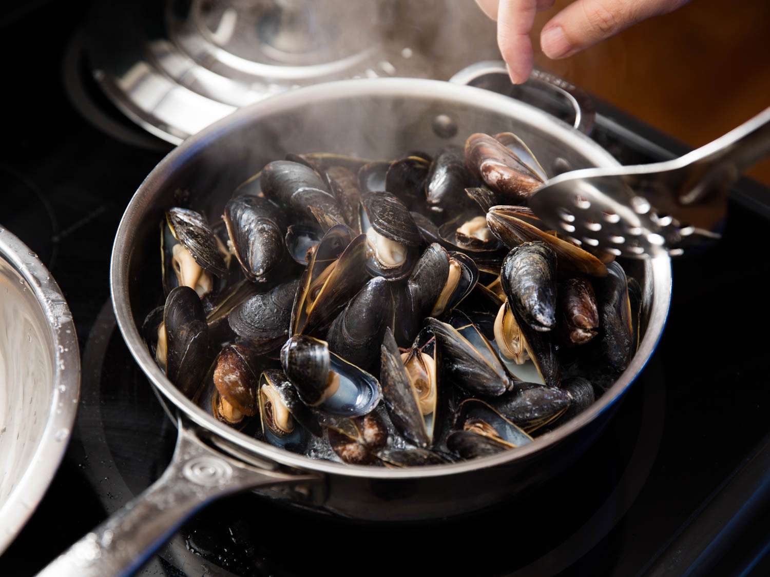 A full skillet of cooking mussels being stirred by a metal spoon.