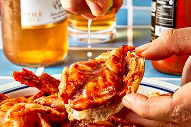 A hand squeezing lime onto a shrimp dish with sauce on a plate beer bottles in the background