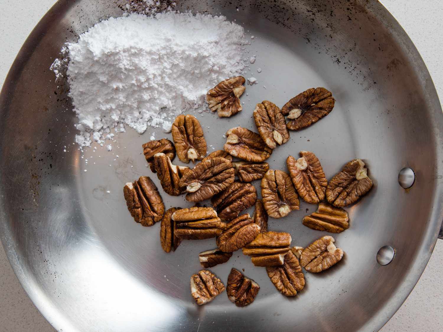 Overhead view of a stainless steel skillet containing a small mound of powdered sugar and raw pecan halves.