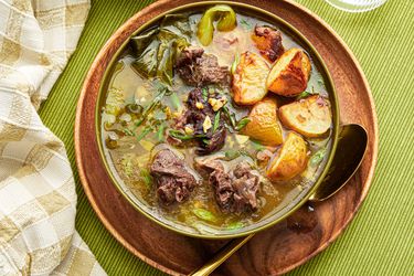 Bowl of Nilagang Beef on a wooden plate, on top of green tablecloth. 2 glasses of water, bowl of soup and napkin on the side. 