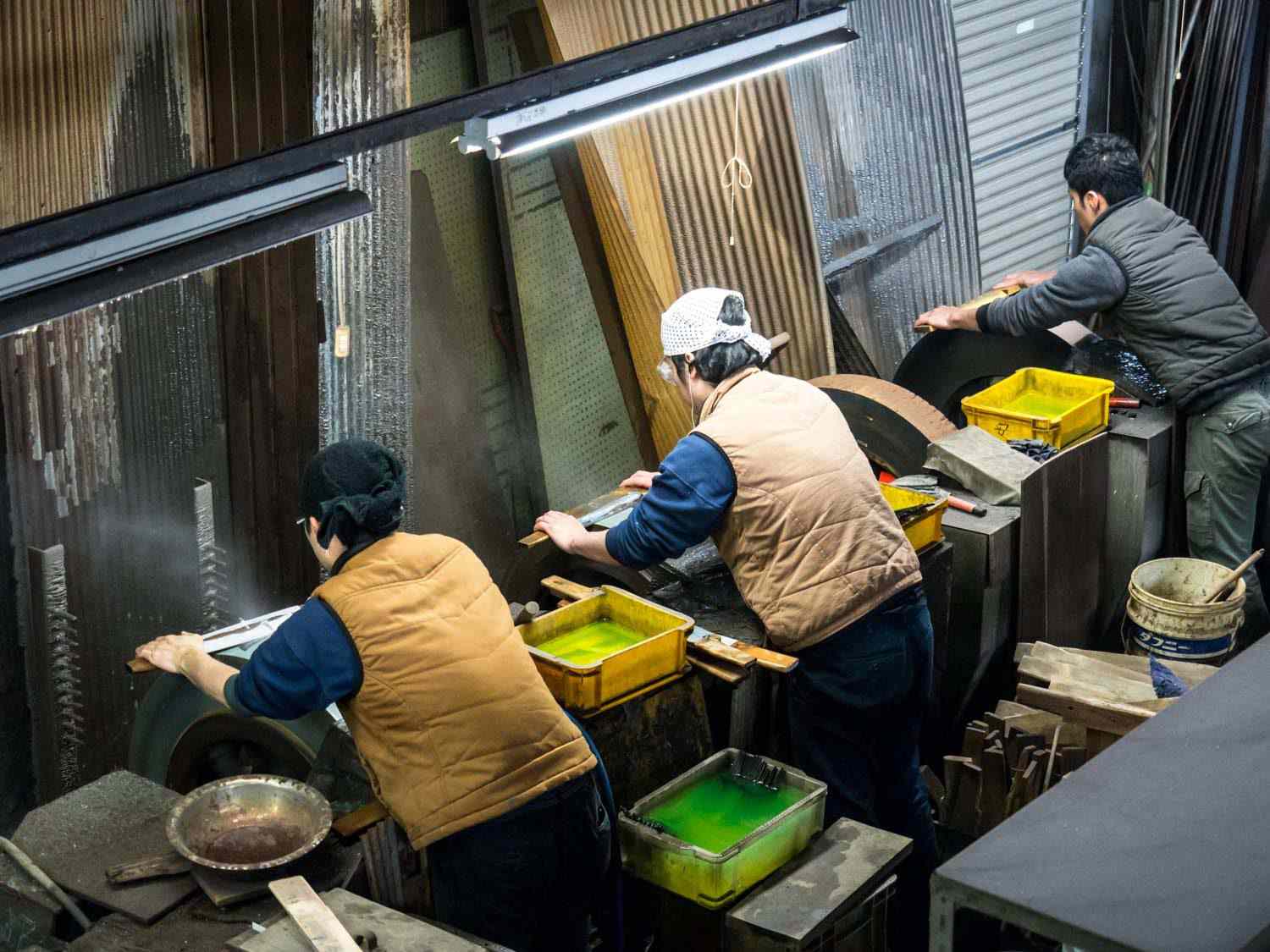 Three bladesmiths hunch over belt grinders as they sharpen finished blades.