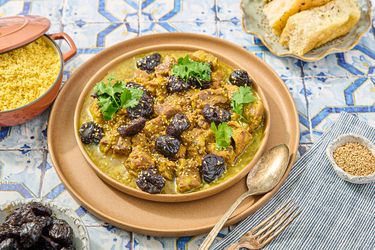 Lamb and prune Tagine with side of couscous, prunes, bread and cutlery on a blue tile surface 