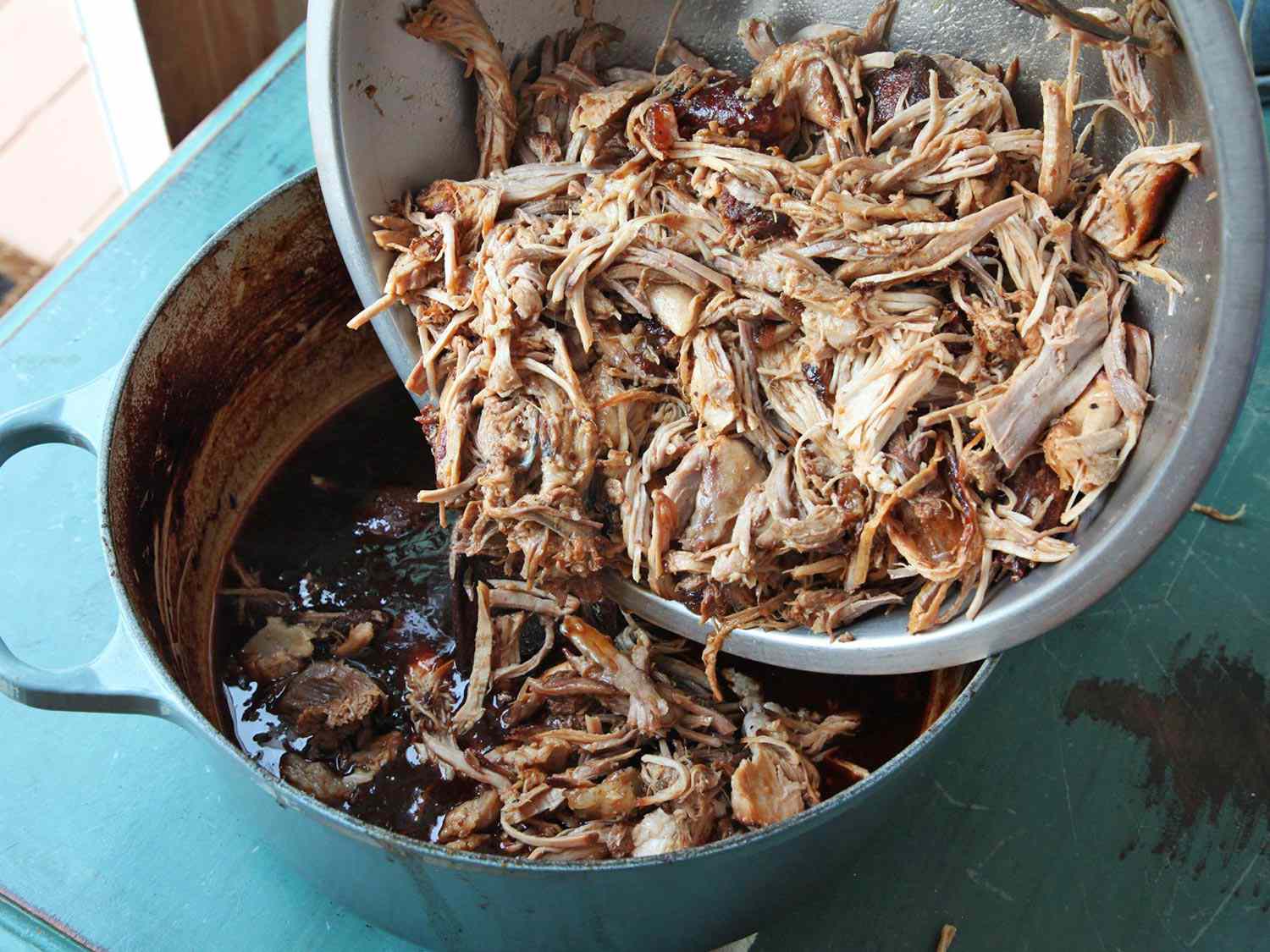 Shredded pork being added to a blue Dutch oven with sauce. 