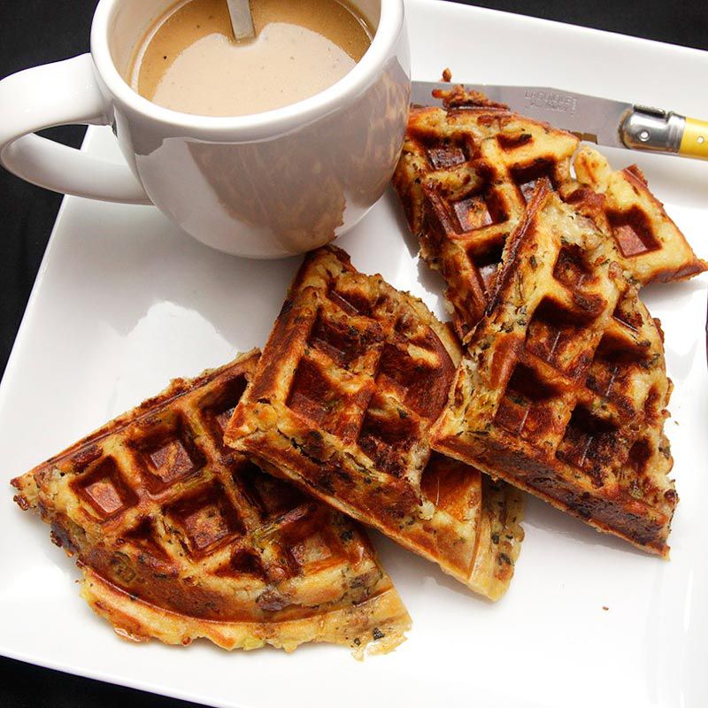 A cooked stuffing waffle quartered, sitting on a white plate next to a knife and a pitcher of gravy syrup.