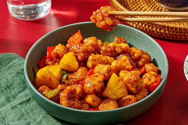 A bowl of Chinese sweet and sour pork with pineapple and vegetables chopsticks holding a piece above the dish
