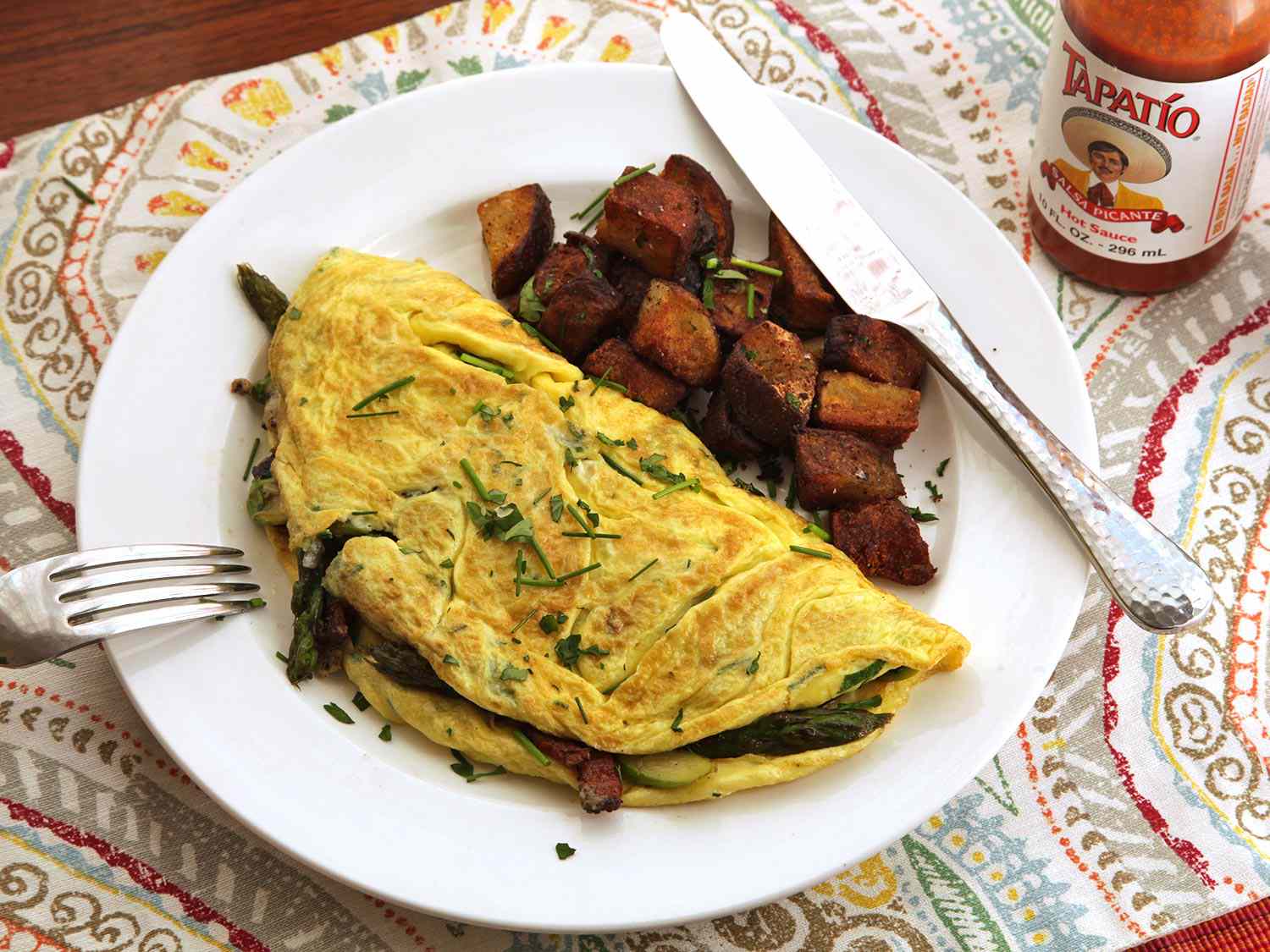 Overhead view of a diner-style asparagus, bacon, and Gruyère omelette for two, served on a white plate with home fries and hot sauce.