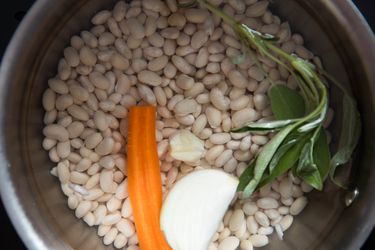 A pot filled with dry beans, a carrot, a garlic clove, onion, and a sprig of fresh sage. 