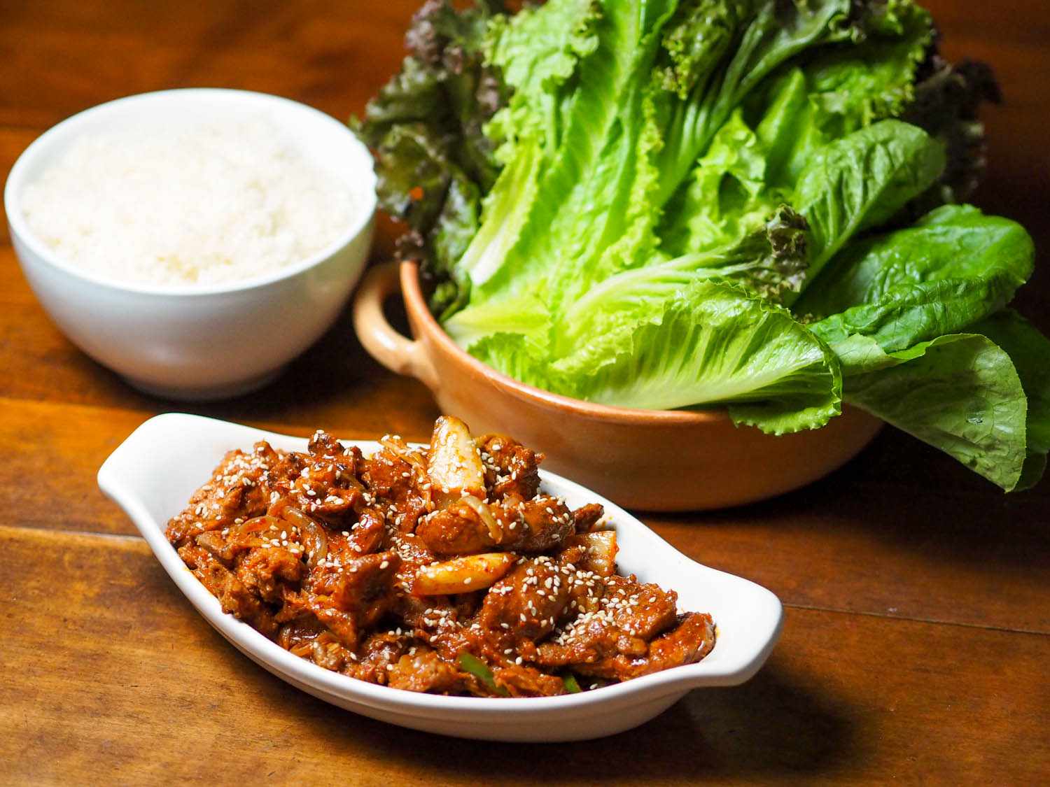 A small serving vessel of jaeyook kimchi bokum, flanked by a bowl of rice and a bowl of whole lettuce leaves.