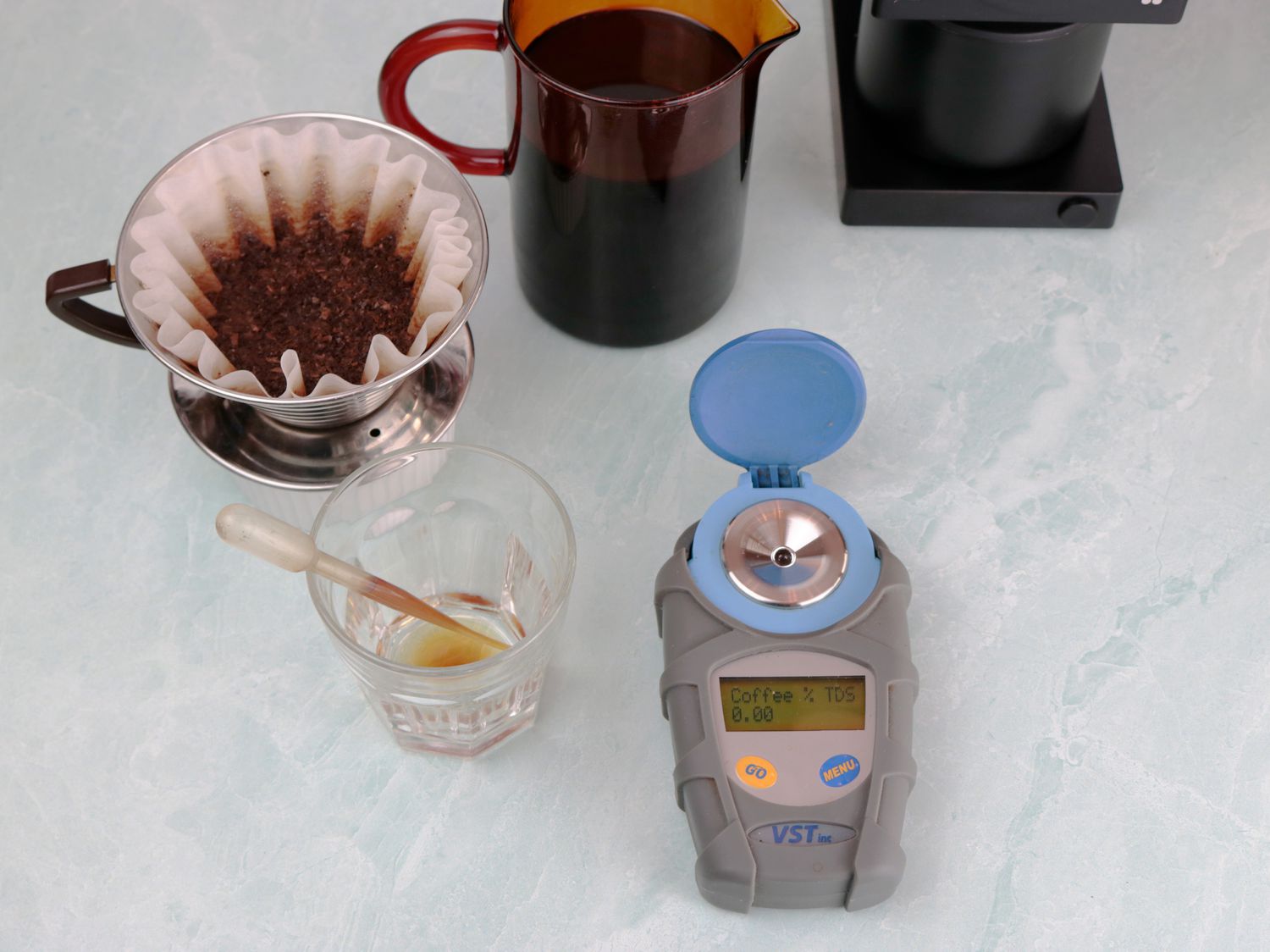 a pourover, coffee carafe, and refractometer on a green background