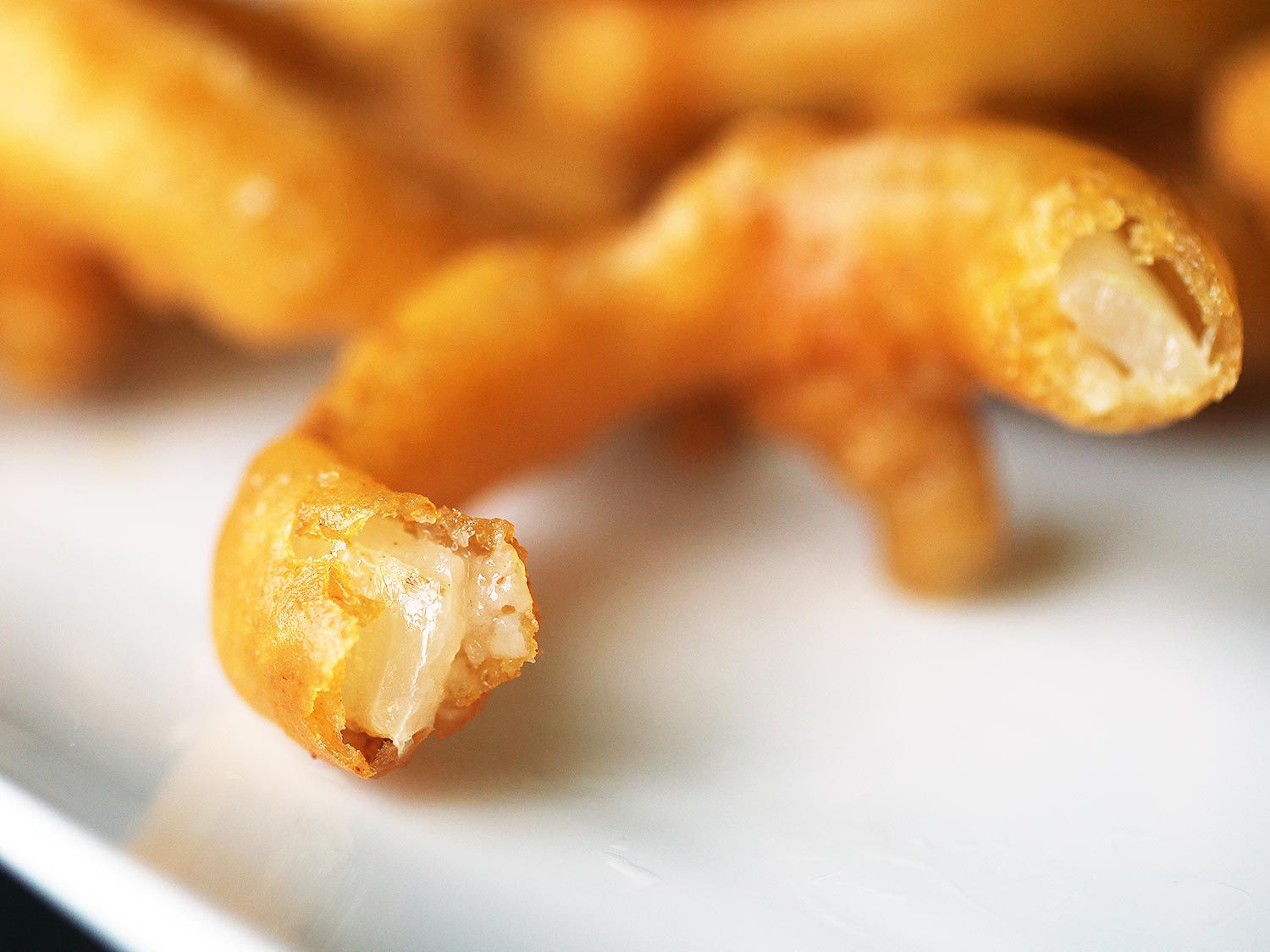Close-up of a bitten onion ring, with a pile of onion rings in the background. The only portion in focus is the bitten ends, where a thin, golden, and crispy shell encases tender, translucent onion flesh.