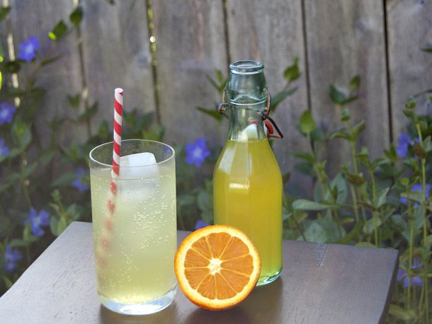 A glass of DIY orange soda with a striped straw next to a bottle of orange concentrate and a sliced orange.