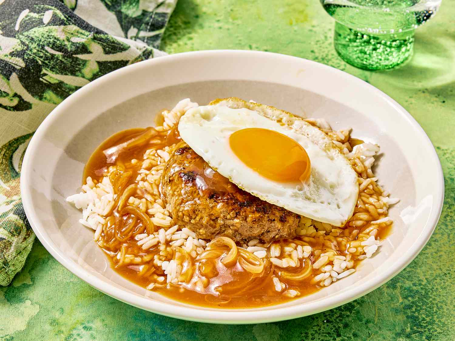 A plate of loco moco, a dish comprising rice, a hamburger patty, gravy, and a fried egg, served on a green patterned tablecloth