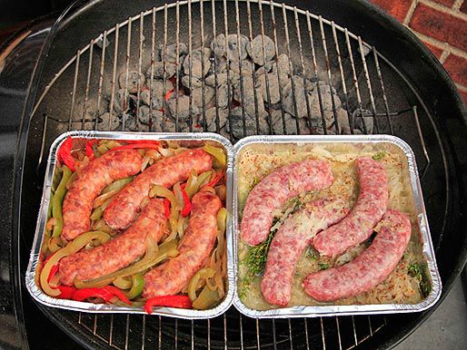 Side-by-side disposable aluminum pans of sausages, one with peppers and onions, one with sauerkraut and mustard, cooking on the grill