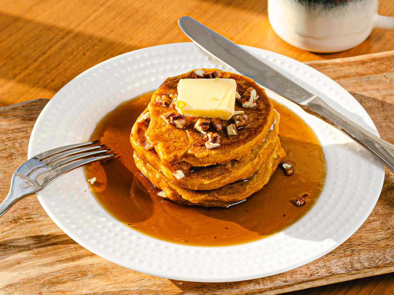 A plate of pancakes with butter syrup and pecans on a wooden surface accompanied by a fork and knife