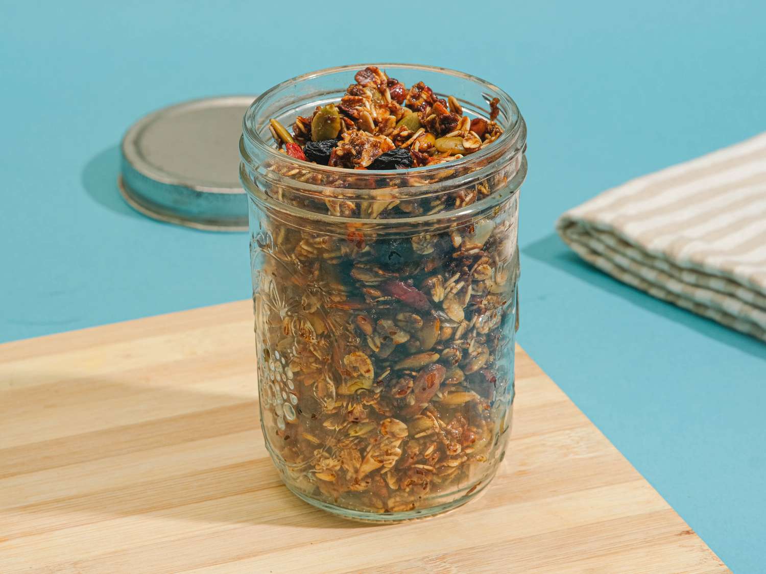 Mason jar with granola, on a wooden board on a blue surface. Napkin and mason jar lid are in the background