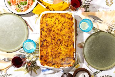 Pastel De Choclo in white dish, on a tabletop with plates, glasses of water and wine, and a salad on the side 