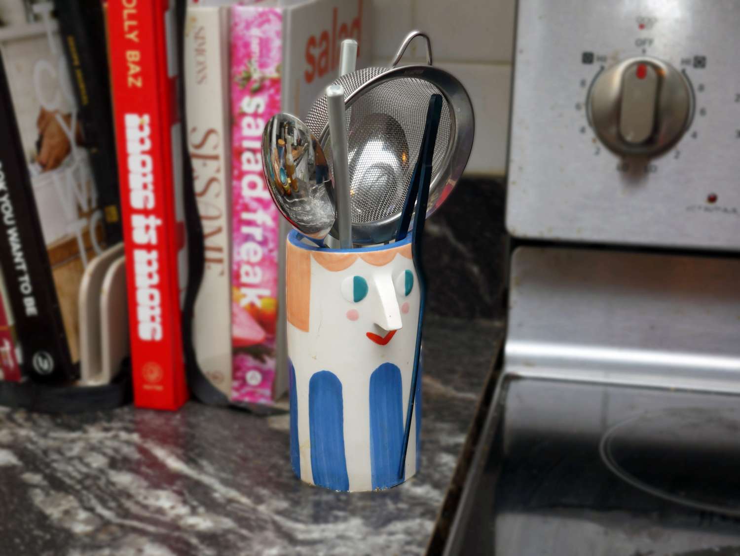Ceramic vase shaped like a face holding kitchen utensils placed on a countertop next to cookbooks and a stove