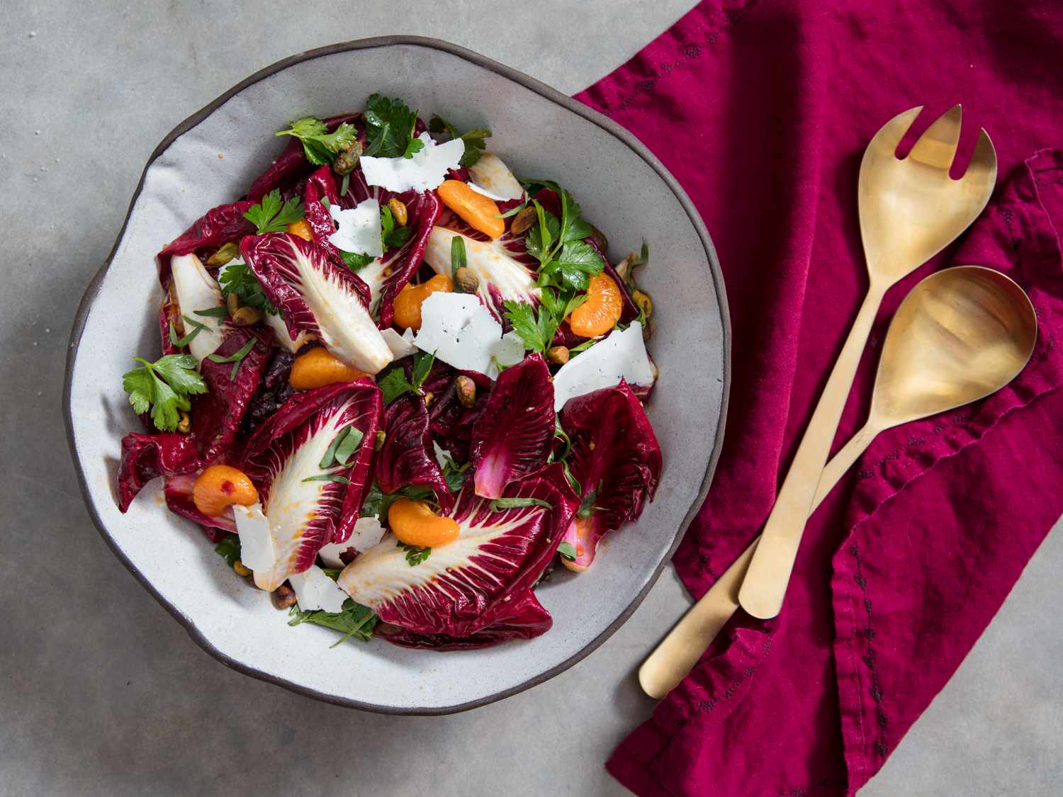 Overhead view of a large ceramic bowl filled with the radicchio salad, dressed and topped with tarragon leaves, pistachios, and thin slices of ricotta salata. Bronze serving implements are close at hand.