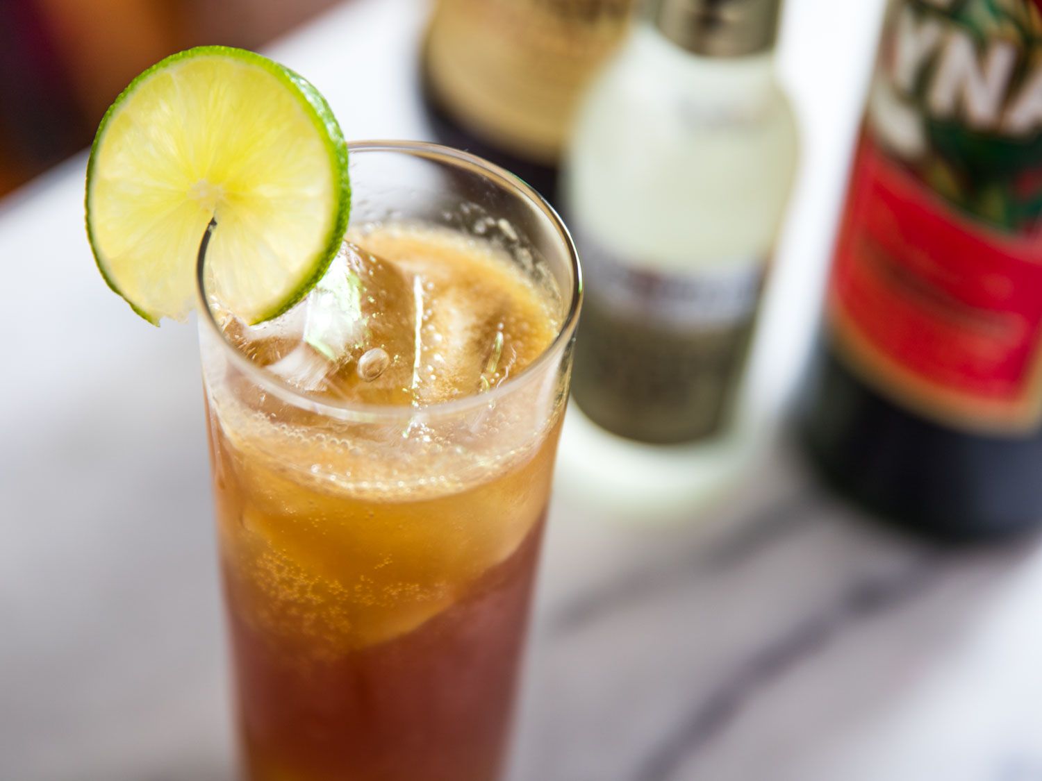 An Italian Buck in a Collins glass garnished with a lime wheel. Ginger beer and Cynar bottles are out of focus in the background.