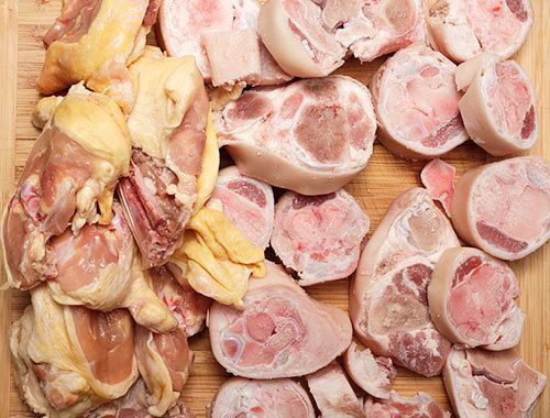 Pieces of chicken carcass and cross-cut pork trotters resting on a wooden cutting board.