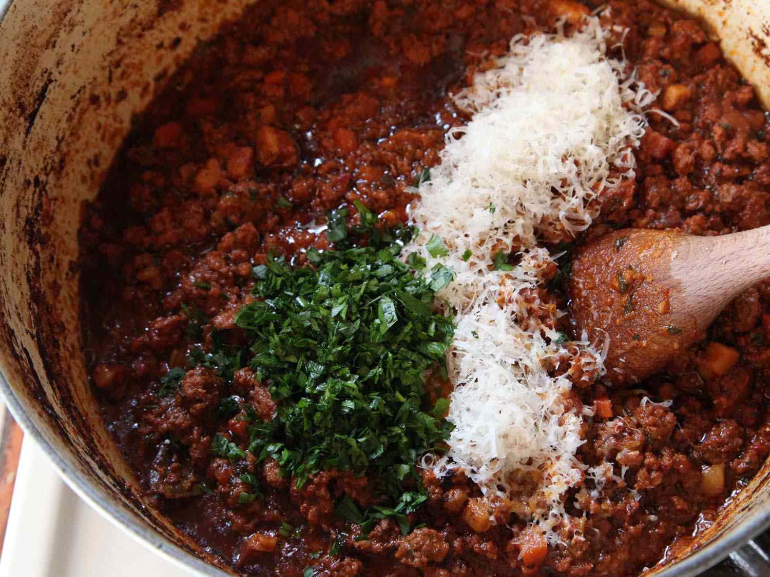 Overhead of parsley and Parmesan cheese being stirred into Bolognese sauce with a wooden spoon.