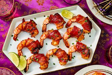 Bacon-wrapped shrimp lined up on a platter, with pink asian inspired fabric, and smaller plates and drinks around the sides