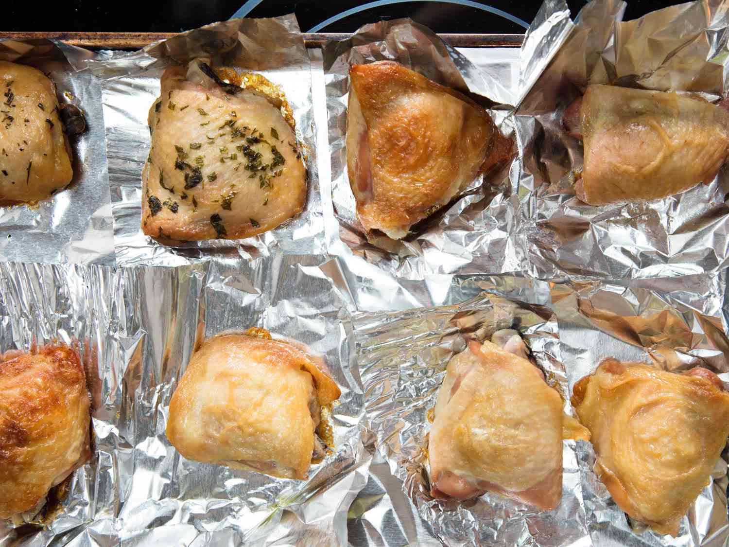 An overhead view of a foil-lined sheet pan full of baked chicken thighs. The thighs look as if they have been prepared for baking dfferently.
