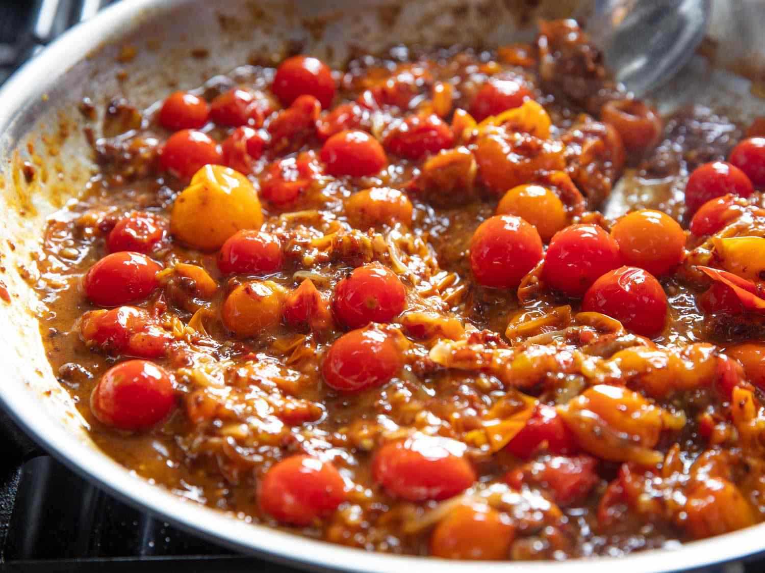Side view of XO sauce being stirred into skillet with burst cherry tomatoes to form a rich sauce.