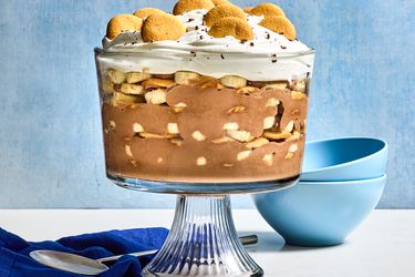 A layered trifle dessert with whipped topping and cookies on top, served in a glass bowl on a stand