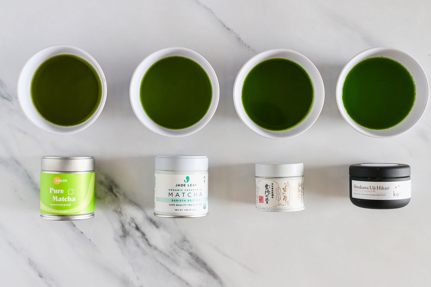 Four bowls of matcha mixed with water beside four tins of matcha.