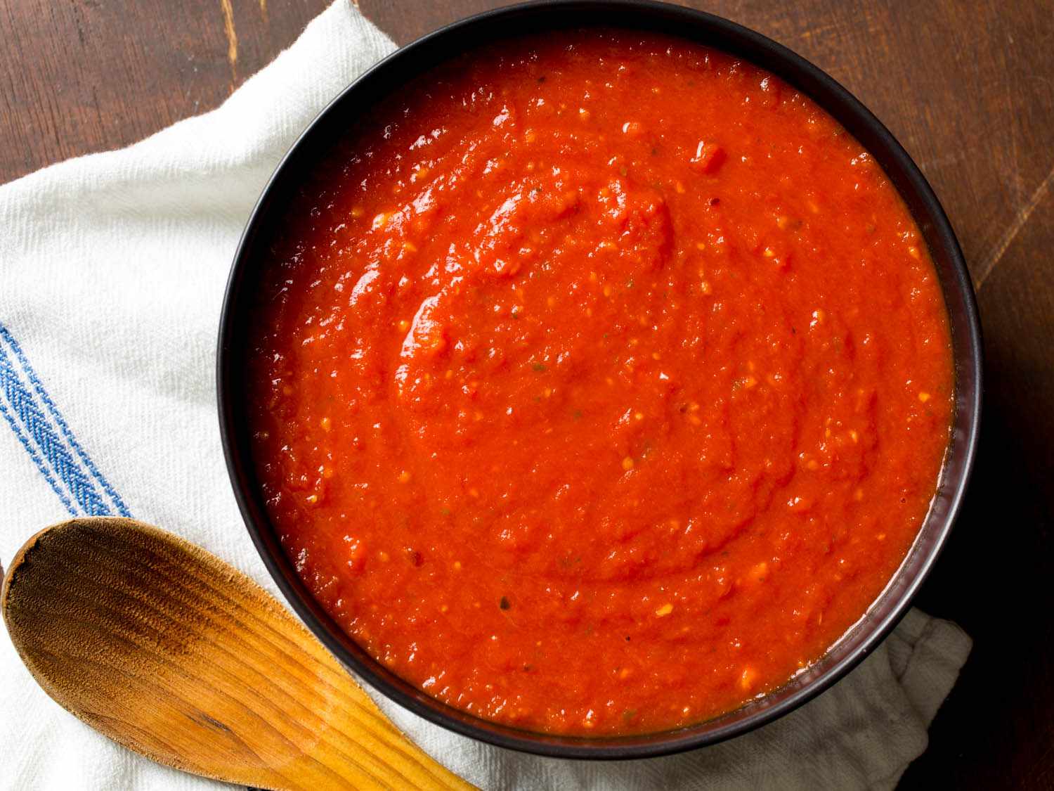 Black bowl filled with quick and easy Italian-American red sauce with towel and wooden spoon on the side.