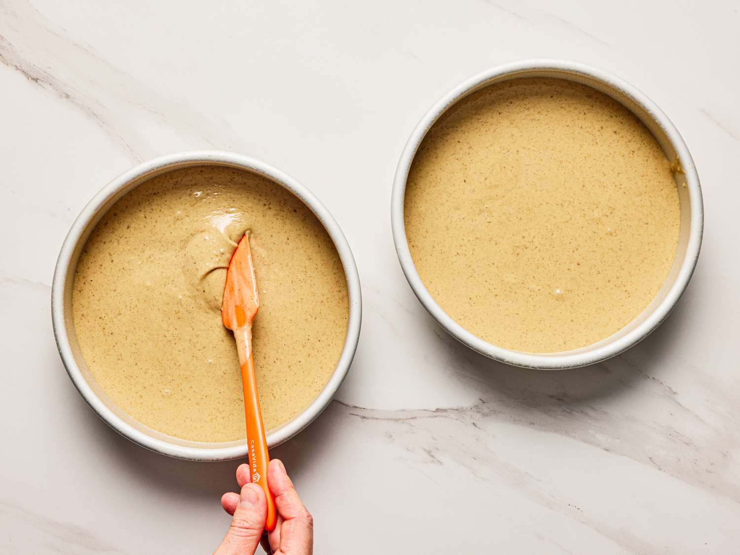 Two round cake pans with batter a hand spreading one with a spatula