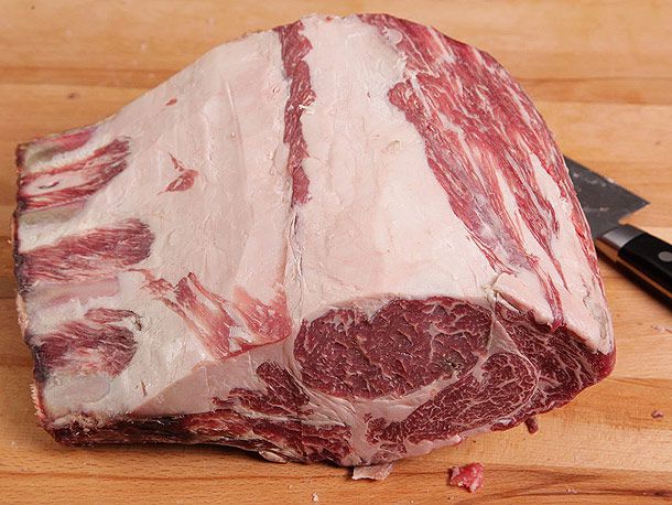 A large, dry-aged prime rib roast that's been trimmed and is ready to cook next to a knife on a wooden cutting board. 