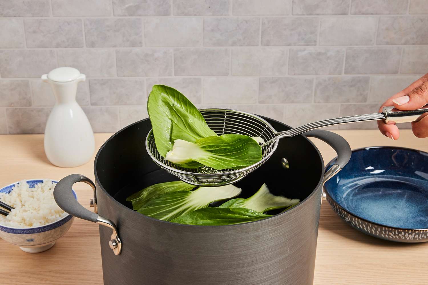 A person uses a metal strainer to lift bok choy leaves out of a black stockpot