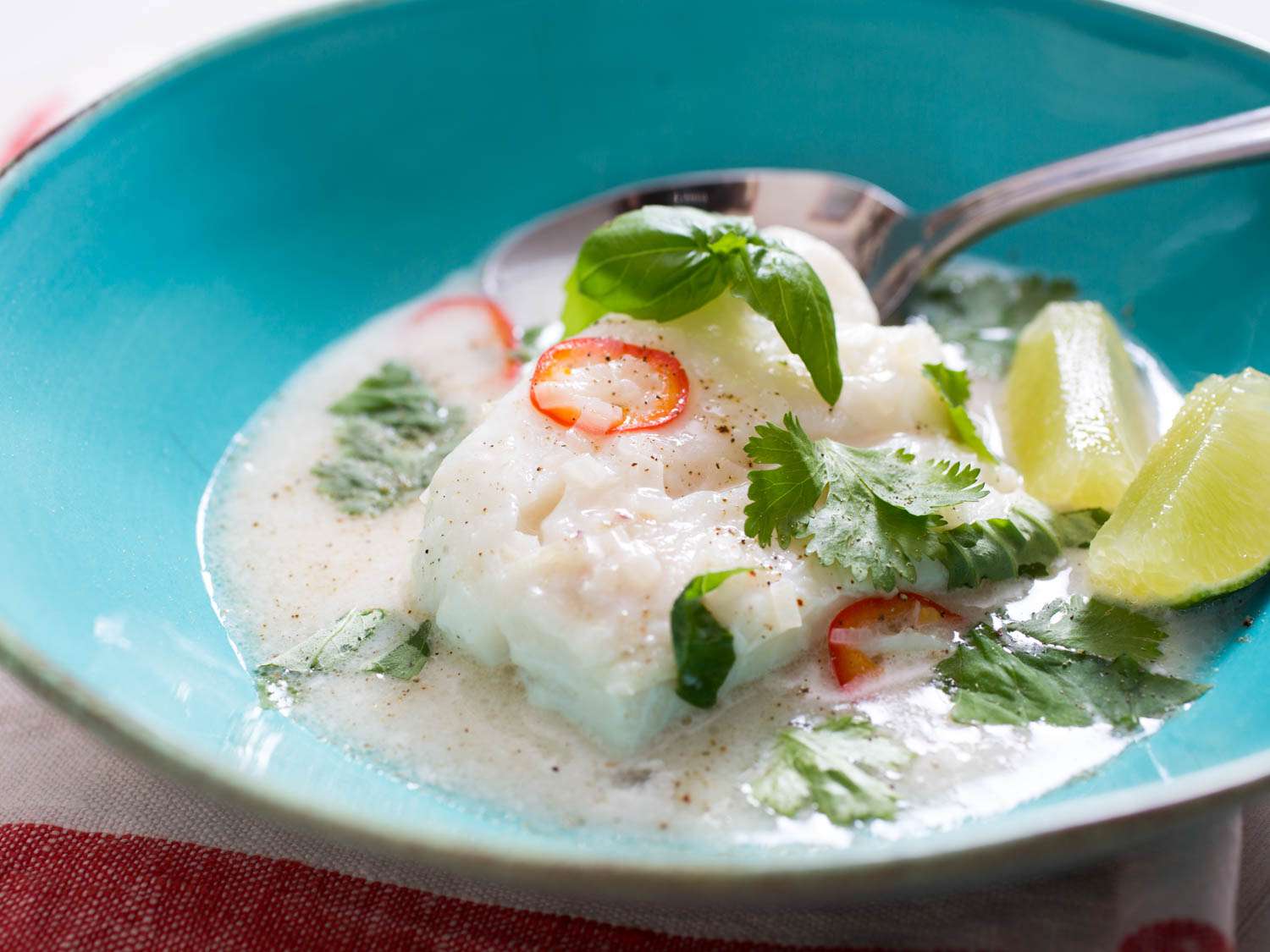 A bowl of cod à la nage with coconut milk, lime, and lemongrass, garnished with cilantro and lime wedges.