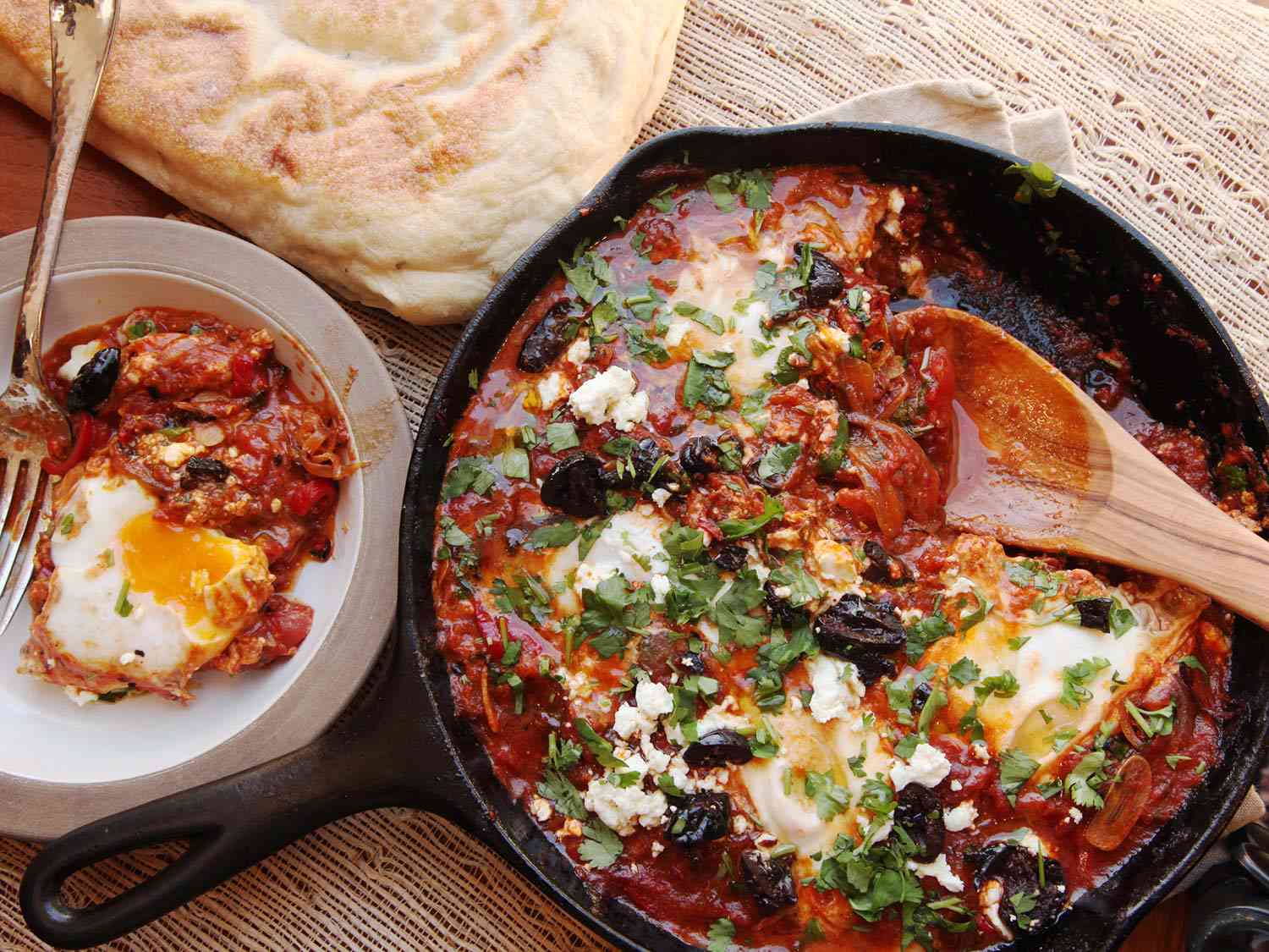 Overhead of a pan with feta-covered shakshuka next to a serving and pita bread.