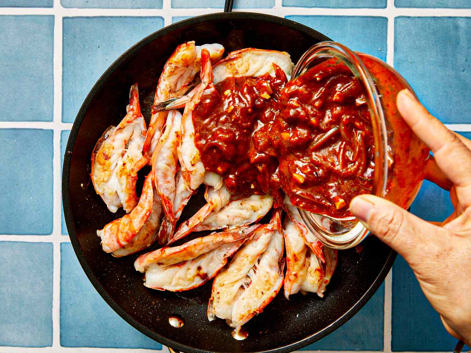 Shrimp in a skillet being topped with a spicy sauce from a jar