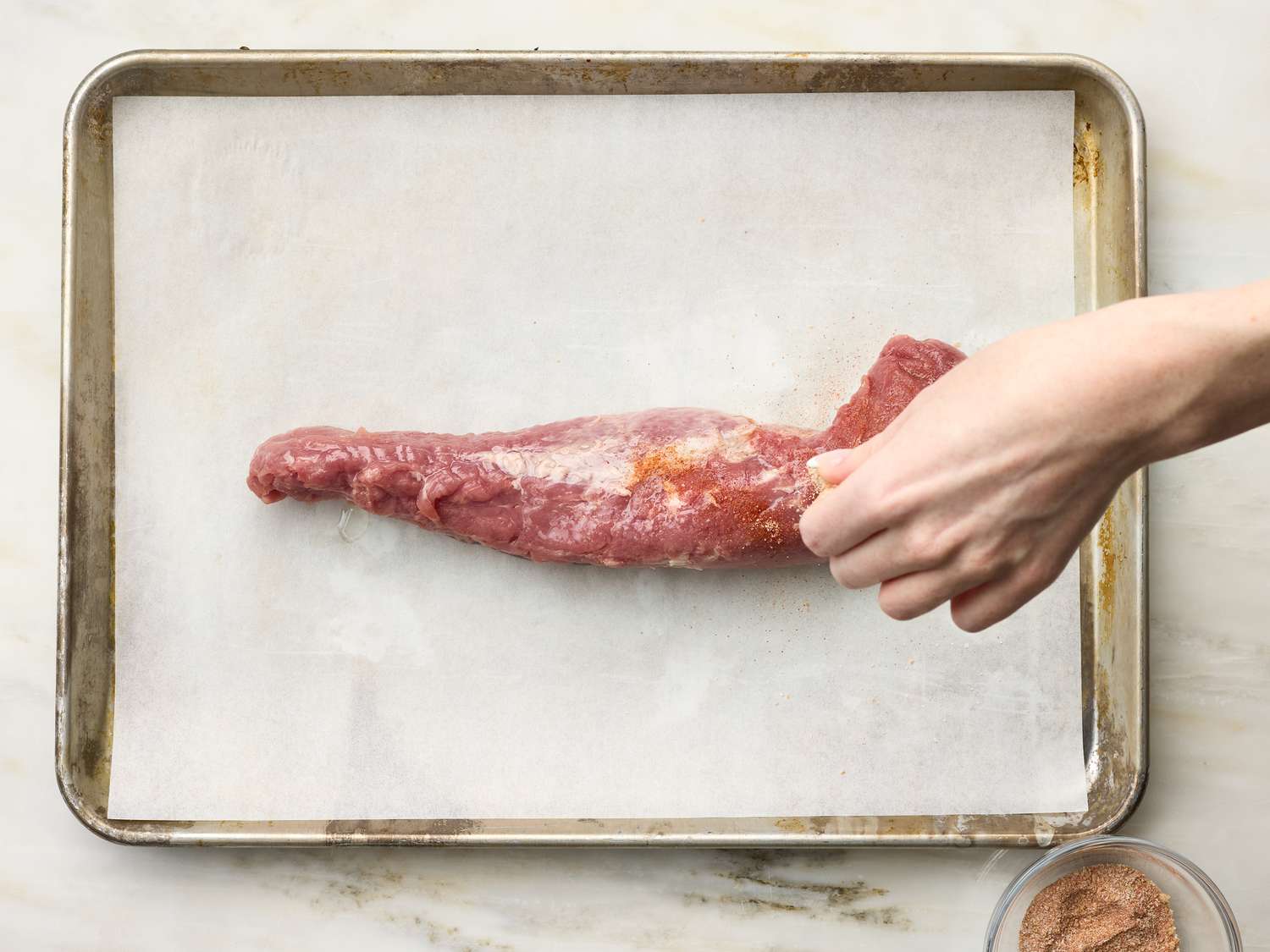 A hand seasoning a pork tenderloin on a baking sheet lined with parchment paper