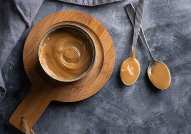 A can of dulce de leche on a wooden trivet with two metal spoons with dulce de leche on them, on a stone background