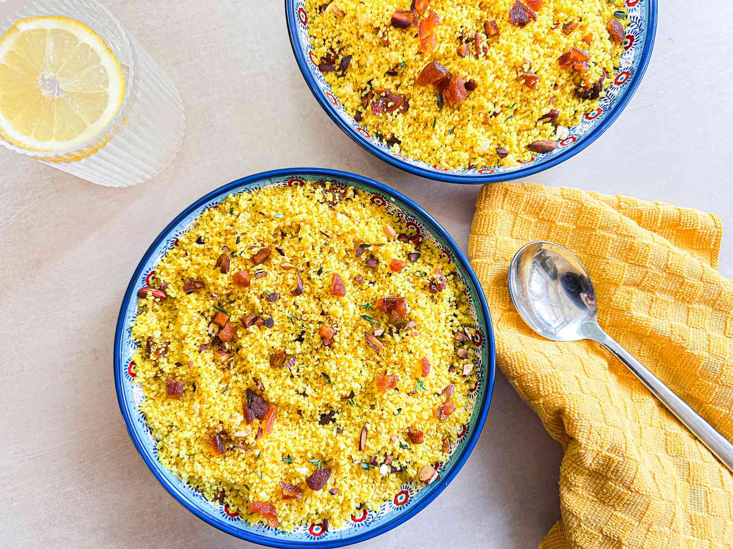 Two bowls of couscous with diced vegetables, a spoon and napkin nearby, and a glass with a lemon slice