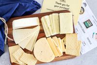 Assorted cheese slices and crackers from Wisconsin Cheese Mart's Wisconsin Cheese of the Month Club on a wooden board with a thank you card and cheese information sheet nearby