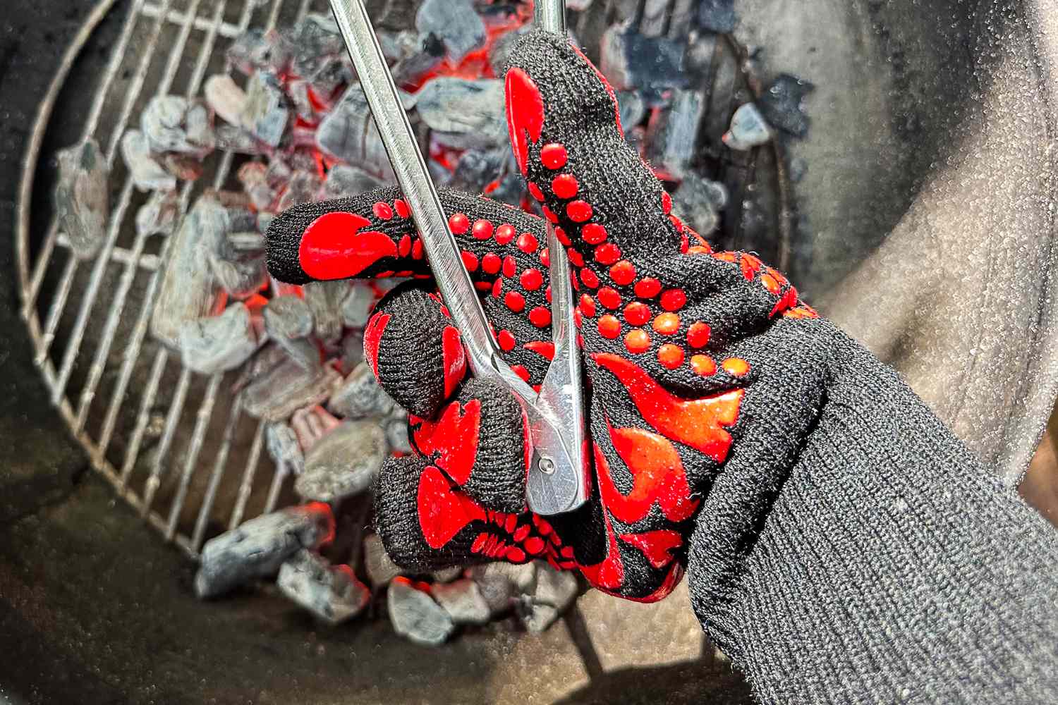 A person holds a pair of tongs over coals while wearing the Comsmart BBQ Gloves