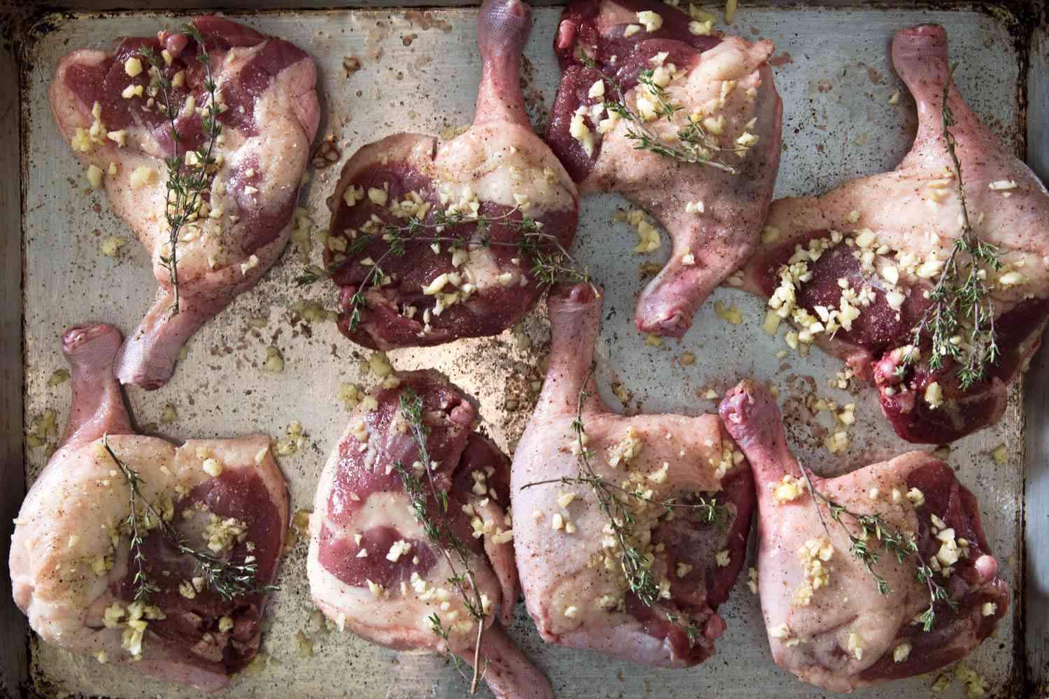 Overhead shot of raw duck legs topped with garlic and thyme sprigs, on a metal baking sheet.