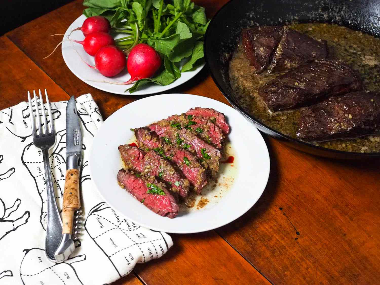 Hanger steaks in a skillet with bagna cauda pan sauce, sliced up and presented on a plate, with a plate of whole top-on red radishes on the side.