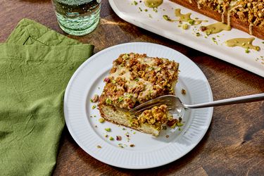 A slice of pistachio coffee cake on a plate with a fork a green napkin and glass of water nearby tray with more cake in the background