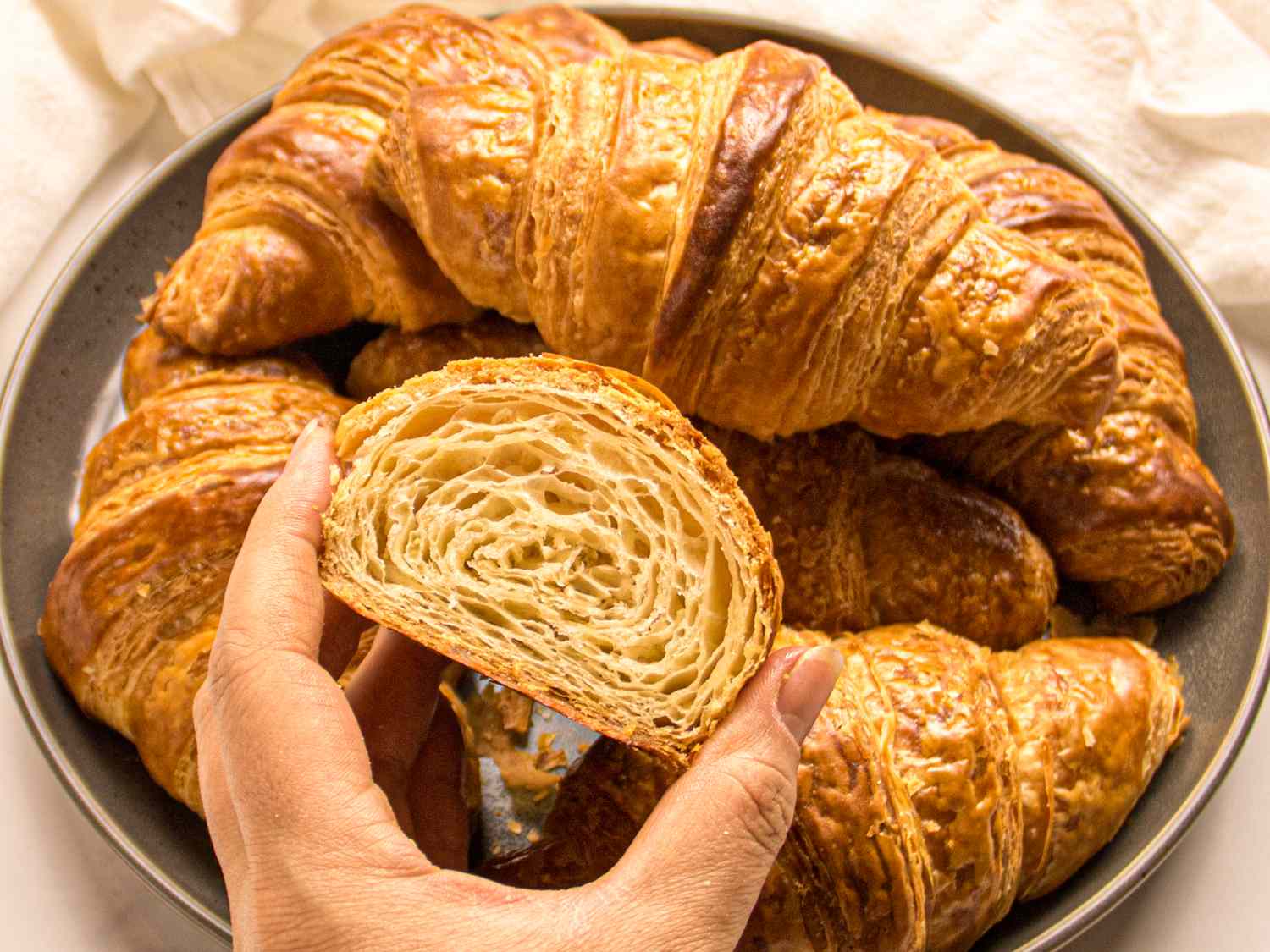 A plate of croissants with a hand holding a halved croissant showing its layers