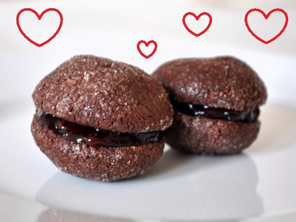 Closeup side view of two chocolate raspberry baci di dama cookies. 4 red, heart-shaped silhouettes have been added to the image.